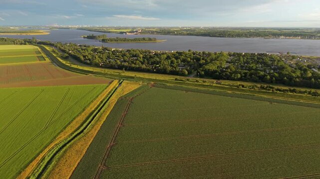 Aerial wide angle panoramic view of lush green agricultural fields and a cozy lakeside village along a wide river under a dramatic cloudy sky in warm evening sunset light