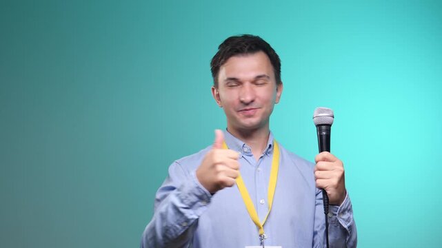 Male presenter with a lanyard and badge giving a thumbs up gesture, indicating approval and success during a public speaking event or conference