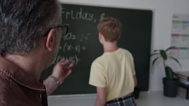 Rack focus shot of mature male teacher explaining math equation to teenager writing on blackboard. Perfect for educational marketing, tutoring services, and school promotional videos
