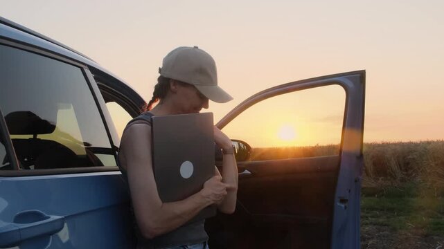 A young farmer woman in work clothes stands with a laptop next to the car in the fields at sunset. She is tired, but satisfied with the result of her work.