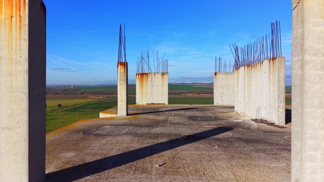 Rows of concrete columns rise above coastal construction site. Overhead perspective reveals rusted rebar tips, glass tower and green farmland beyond.