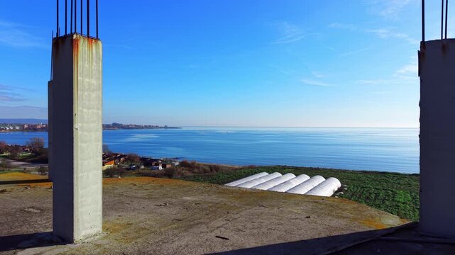 Rooftop view of greenhouse tunnels and coastline, Pomorie, Bulgaria. Overhead angle captures white polytunnels, cabins, farmland and calm turquoise sea. Aerial view of construction site.