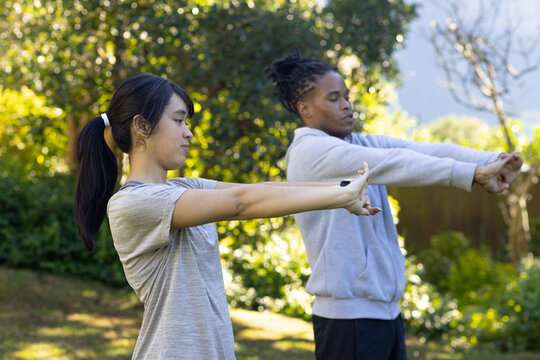 African American man and Asian woman stretching arms in sunny yard wearing T-shirt and watch