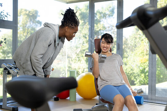 Male trainer leaning forward coaching female client doing bicep curl on gym bench with dumbbells