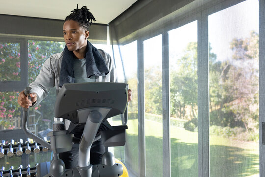 African American man exercising on elliptical in home gym in gray hoodie towel weights windows