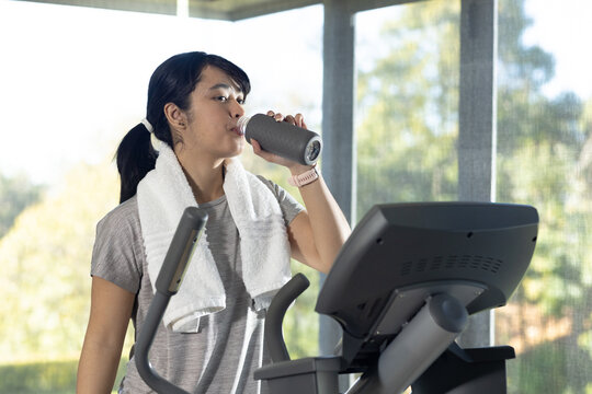 Asian woman standing at cardio machine in gym sipping from textured bottle, towel and watch visible