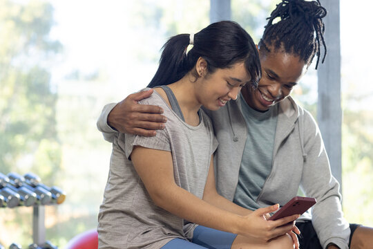 African American man and Asian woman sitting on bench in gym, holding smartphone near dumbbell rack