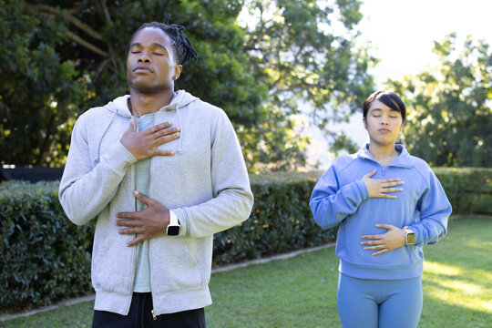 Diverse couple practicing breathing with eyes closed on grass by hedge wearing fitness watches