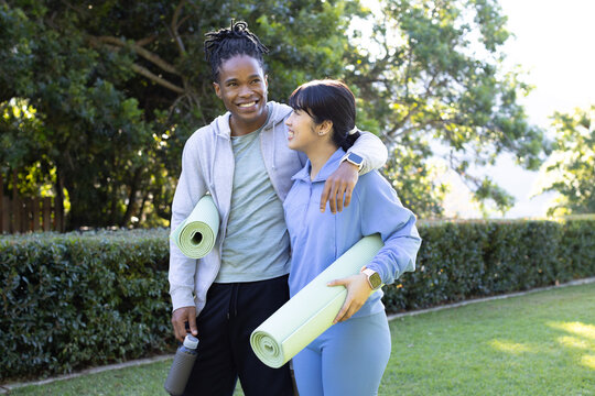 Diverse couple wearing gym gear on lawn holding rolled mats, one with bottle, other with smartwatch