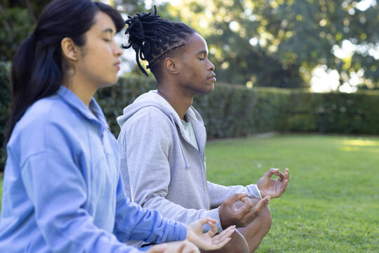 African American man and Asian woman meditating on grassy park lawn wearing hoodie, zip jacket