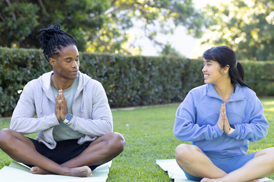 Diverse couple sitting cross-legged on park lawn pressing hands at chest on mats, in sportswear