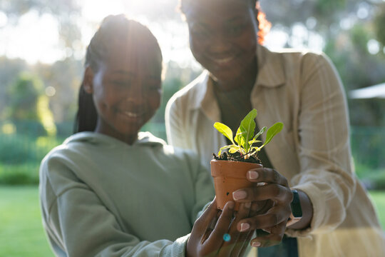 African American mother and youth daughter holding clay pot with seedling, ring and watch in garden