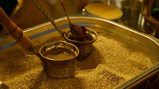 Ground coffee is added into copper cezve during traditional sand brewing process. Street food vendor prepares authentic Turkish coffee at outdoor food stall.