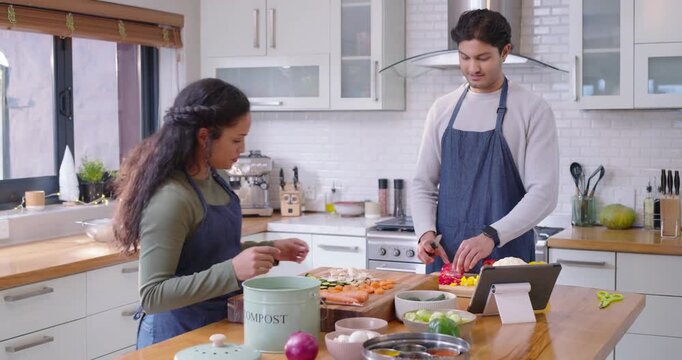 African American couple wearing aprons chopping vegs at island following tablet recipe, composting