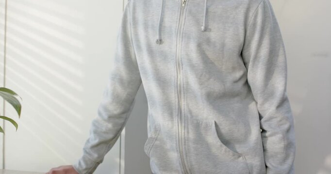 Male wearing gray hoodie placing hand on counter at window with blinds, plant, turning left smiling