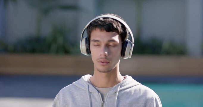 African American man standing eyes closed, listening on headphones, wearing gray hoodie by pool