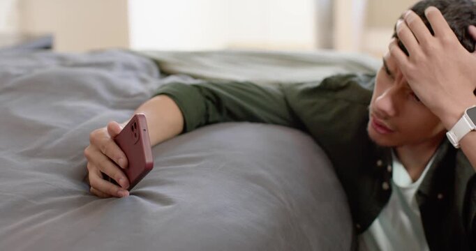 African American man in 20s noticing maroon phone on bed, reaching, checking messages, rubbing brow