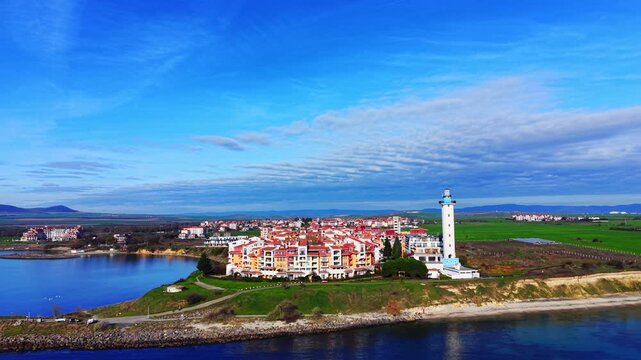 Coastal resort with lighthouse overlooks bay, Pomorie, Bulgaria. Aerial drone view shows colorful buildings, greenery, cliffside path and calm blue water.