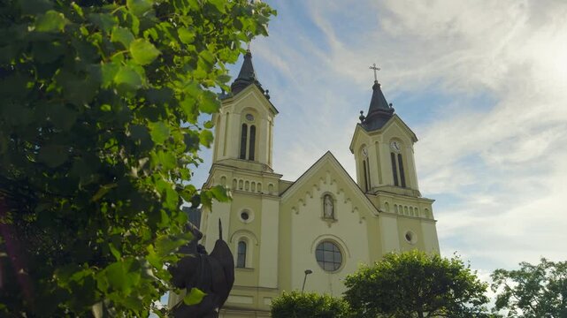 Slow push-in toward historic twin-tower church framed by lush green trees on a sunny day - Parish Church of the Transfiguration, Sanok, Poland.
