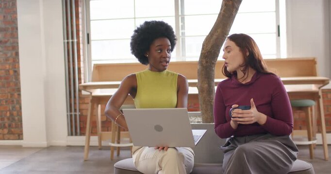 Diverse female coworkers with left woman opening laptop, collaborating and holding blue mug at work