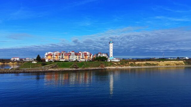 Waterfront view of Pomorie lighthouse and resort buildings, Bulgaria. Calm reflection, cliff shoreline and cloudy blue sky.