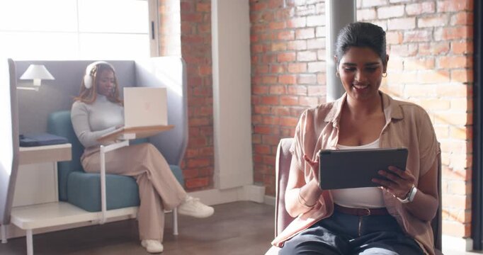 Diverse female team working at lounge, camera panning, pod user typing laptop, peer tapping tablet