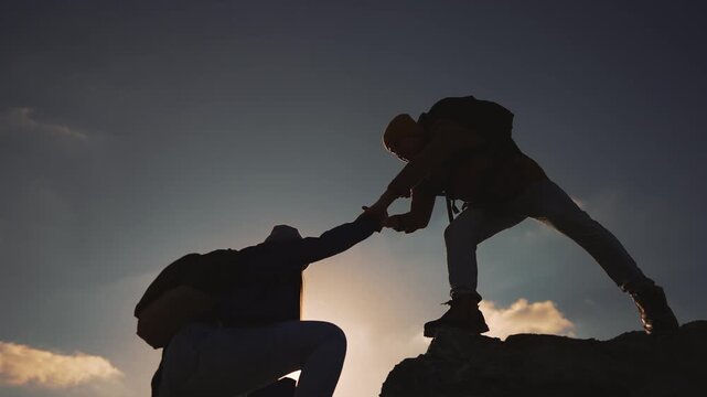 Silhouette of two hikers helping each other climb steep rocky mountain terrain at sunset. One person extends hand to assist partner up challenging slope during outdoor adventure expedition.