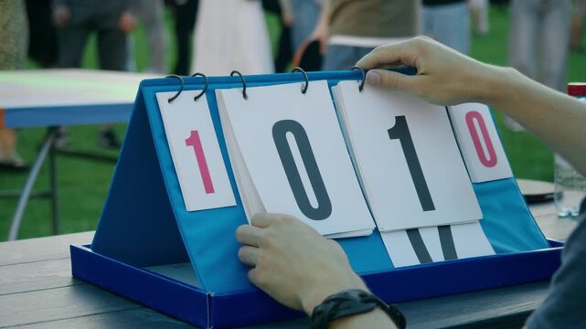 Scorekeeper flips scoreboard card after first point during amateur outdoor table tennis tournament at summer party. Manual scoreboard shows player taking early lead as friends watch exciting ping pong