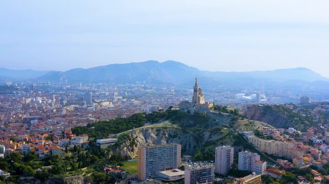 Aerial view of Marseille, France, with the Notre Dame de la Garde basilica and its golden statue on the hilltop, the Stade Velodrome roof, residential districts and Provence mountains.