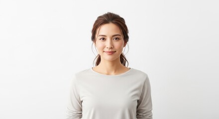 Obraz premium A young woman with long brown hair, wearing a light gray long-sleeved top, standing against a plain white background.