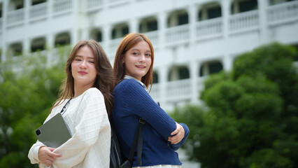 Fototapeta premium Two confident female students stand together on a university campus holding a tablet, reflecting friendship, academic success, and a modern college lifestyle outdoors.