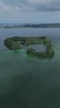 Aerial drone footage circling De Schelp island in Gooimeer, Netherlands. Small green recreational island surrounded by open lake water, with trees, sheltered lagoon, cloudy sky and distant shoreline.