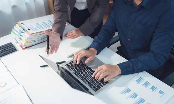 Business Meeting and Analysis: Two professionals collaborate over a laptop, surrounded by documents and charts, analyzing data and planning strategies in a modern office setting.