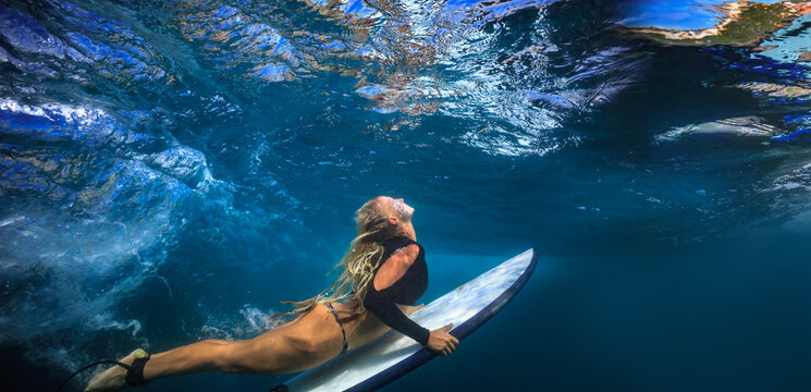 Underwater view of blonde female surfer paddling towards surface light