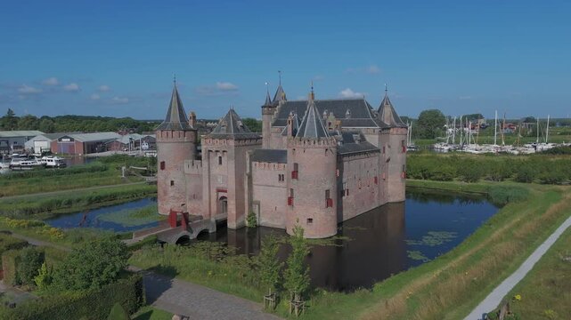 Aerial drone footage circling a medieval brick castle surrounded by a moat and green landscape. Short orbit shot showing the historic fortress, towers and nearby marina under a clear blue sky.