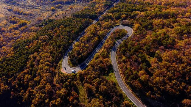 Overhead perspective of hairpin mountain road. Switchback curve through dense autumn woodland with a van navigating the bend.