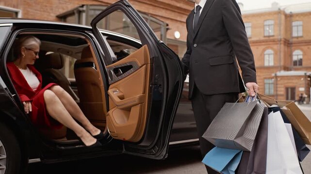 Senior Caucasian woman getting into luxury car while male chauffeur holding shopping bags closes door. Ideal for high-end retail, wealth management, and premium concierge service commercials