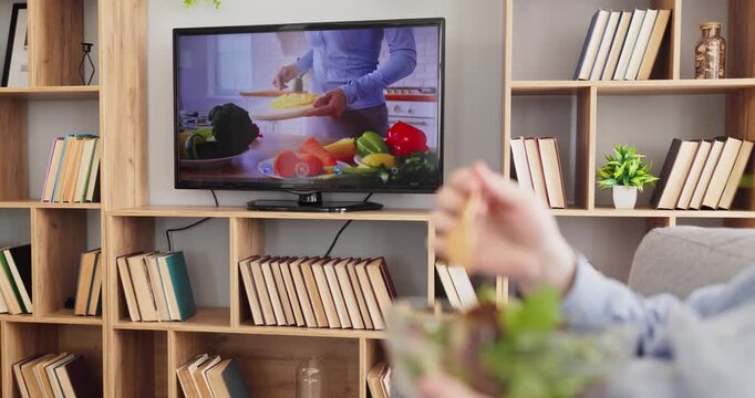 Man at home watching television eating salad. A male adult enjoys a fresh diet as a cooking show inspires health, nutrition tips, and wellness at home. Focus on healthy lifestyle and nutrition.