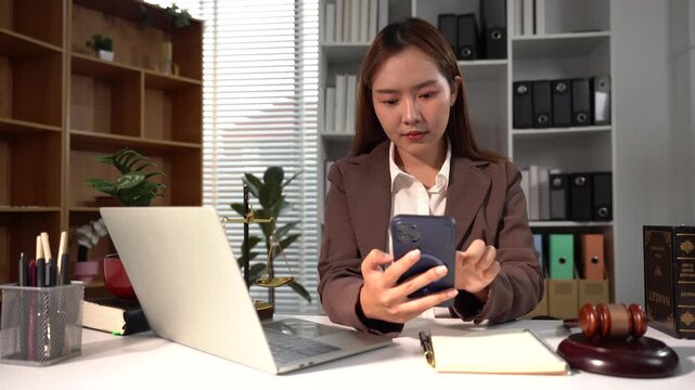 Lawyer Working on Phone: A focused lawyer in a professional setting uses her smartphone while sitting at a desk with a laptop and legal gavel, symbolizing modern legal practice.