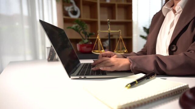 Lawyer Working on Laptop: A legal professional types on a laptop in an office, with scales of justice and law books visible in the background, symbolizing law and justice.