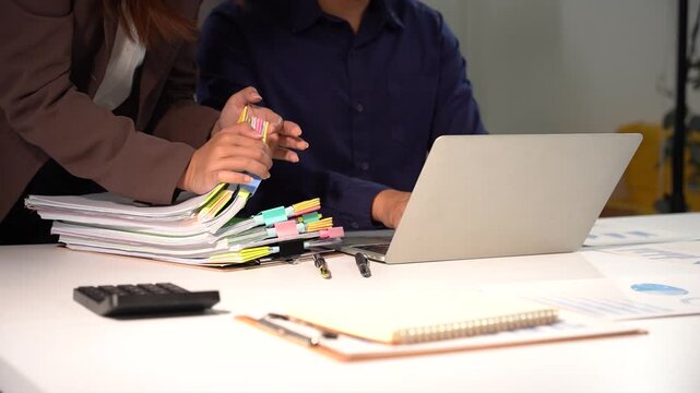 Collaborative Effort: Two individuals are seen working together in an office, one pointing to documents while the other uses a laptop, signifying teamwork and shared workload.