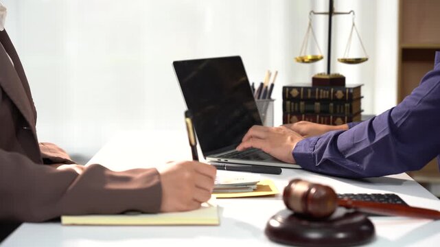 Legal Consultation: Two individuals engage in a professional discussion in an office setting, with a laptop, legal books, and scales of justice visible, signifying legal proceedings or consultation.