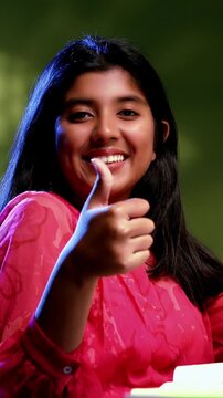 Indian Asian studious teenage beautiful girl sitting at a study table in a cozy lavish home, showing a confident success gesture while studying, writing, demonstrating focus, determination, excellence