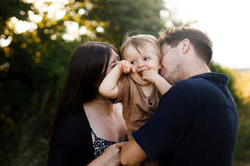 Fototapeta premium Beautiful joyful mother and father hugging their smiling son in summer countryside. Happy family kissing and cuddling on background of sunflowers sunny field. Happiness and togetherness