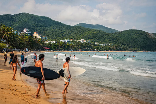 Surfers carrying boards through shallow waves on a tropical Hainan beach.generative AI