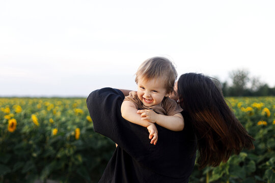 Happy mother playing with son in sunny summer field. Stylish mother holding baby boy, having fun and smiling on background of sunflowers. Family leisure with a child