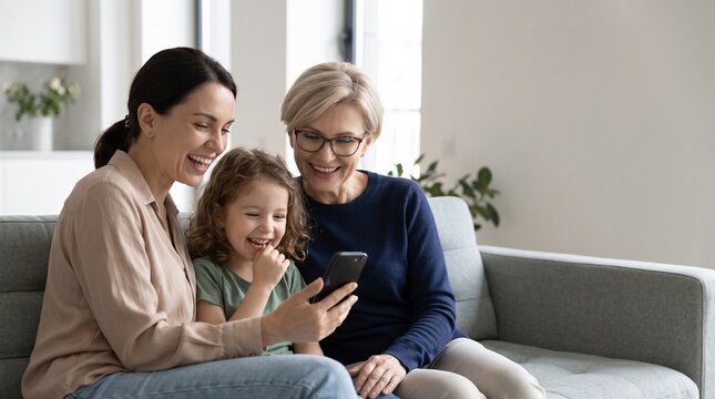 Happy three generations of family looking at smartphone at home. Smiling mother curly hair child girl and grandmother with glasses watching funny online content on cozy sofa in living room.