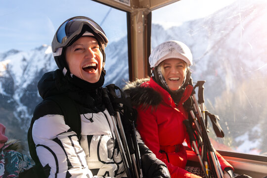Two female skiers laugh together inside mountain gondola, holding poles beside snowy alpine scenery. Joyful winter moment captures friendship, ski fun, happiness, travel, and holiday energy