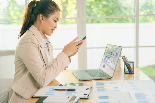 A smiling Asian businesswoman works with financial data charts and a smartphone in the office, analyzing business data graphs.