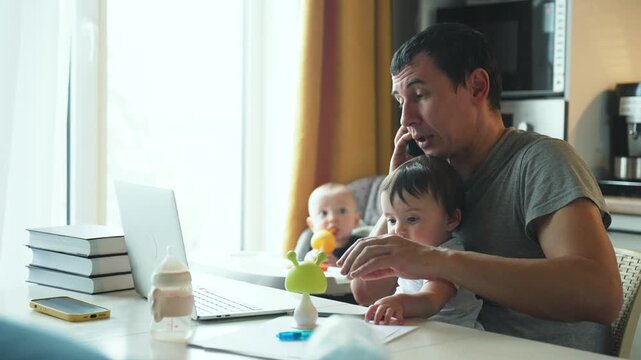 Busy father multitasking on laptop while holding his toddler son at kitchen table. Dad works from home managing two young children. Parent balancing remote work and childcare responsibilities.